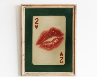 May include: Framed artwork featuring a playing card with a red lip print. The card is the two of hearts, with the number 2 and a heart symbol in each corner. The background is a dark green, and the frame is a light wood color.