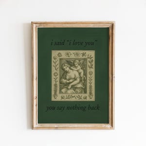 Op de afbeelding: Ingelijste kunstdruk met een donkergroene achtergrond en een vintage illustratie van een vrouw en een engel. De tekst "i said 'i love you'" en "you say nothing back" zijn op het kunstwerk gedrukt. De lijst is lichtbruin.