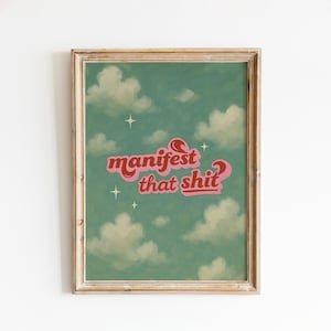 Può includere: Stampa artistica incorniciata con un cielo turchese, nuvole bianche e quattro stelle. Il testo "manifest that shit" è in un font retrò con colori rosa e rosso. La cornice è di colore marrone chiaro.