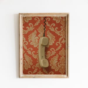 May include: A framed artwork featuring a vintage telephone receiver hanging against a red and gold damask wallpaper background. The frame is a light, neutral colour, and the phone is a muted green.