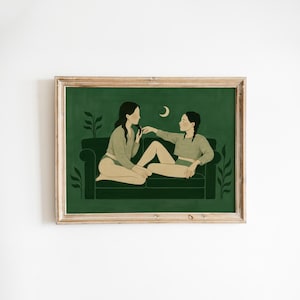 May include: A framed art print showing two figures on a dark green sofa. The figures are in soft colors, with one seated and the other reclining. A crescent moon is in the background. The artwork is framed in a light brown wooden frame.