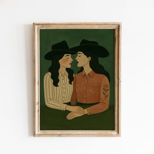 May include: A framed art print featuring two figures in cowboy hats, facing each other. One figure wears a striped shirt, the other a rust-colored shirt with a rose tattoo. The background is a dark green, and the frame is a light wood color.
