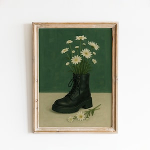 May include: A framed art print featuring a black combat boot filled with white daisies against a green backdrop. The boot has a chunky sole and is laced up. A few daisies lay on the surface in front of the boot. The frame is a light brown color.