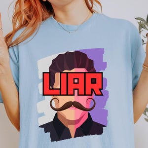 May include: A light blue t-shirt featuring a graphic print of a stylized person with dark hair and a prominent handlebar mustache. The word LIAR is printed in bold red letters across the eyes. The background of the graphic is a split white and purple.