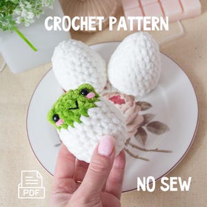 May include: A crocheted frog emerging from a white crocheted egg, with two more white crocheted eggs on a floral plate. The frog is green with black eyes and pink cheeks. The text "CROCHET PATTERN" and "NO SEW" are visible.