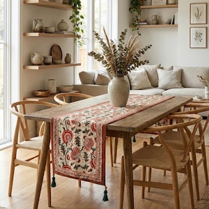 Puede incluir: Un comedor con una mesa de madera adornada con un camino de mesa floral. Sillas de madera con asientos tejidos rodean la mesa. Un jarrón con flores secas está sobre la mesa. Estantes con objetos decorativos y arte enmarcados son visibles.