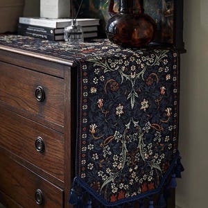 May include: A dark blue floral patterned table runner with a decorative tassel trim. The runner is placed on a dark wood dresser. A vase of white flowers sits on top of the dresser.