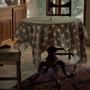 May include: A round tablecloth with a dark green background and a repeating pattern of gold starbursts. The tablecloth is draped over a dark wooden table with a decorative base. A small glass sits on the table. The scene is set in a room with wooden furniture.