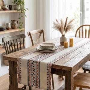 Può includere: Un tavolo da pranzo rustico in legno è apparecchiato con un runner color crema con un motivo geometrico blu navy e arancione bruciato. Piatti, candele e un vaso di fiori secchi creano un'atmosfera calda e invitante.
