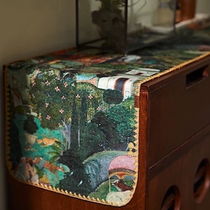 May include: A table runner with a detailed landscape design featuring trees, water, and figures. The runner has a yellow stitched border and is placed on a dark brown wooden dresser. The artwork is predominantly green, blue, and brown.