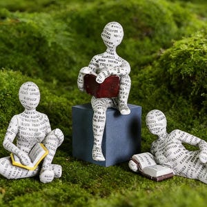 Peut inclure: Trois figurines originales de personnes lisant des livres. Les figurines sont recouvertes de texte et sont dans diverses poses : assises, allongées et perchées sur un cube bleu. Les livres sont dans des tons de rouge, jaune et marron.