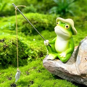 Puede incluir: Una figura de rana verde con sombrero de paja, pescando con una caña plateada y un pequeño señuelo de pez. La rana está sentada en una roca en un entorno cubierto de musgo. Es un artículo decorativo.