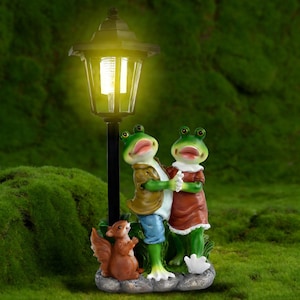Puede incluir: Una farola de jardín caprichosa con dos figuras de ranas verdes con ropa formal, tomadas de la mano. Una figura de ardilla marrón está en la base. La lámpara tiene un poste negro y una luz amarilla.