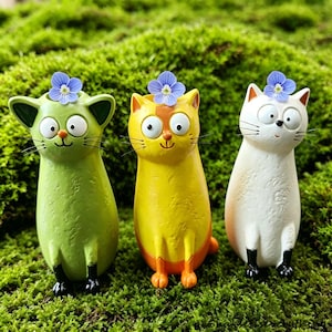 May include: Three cat-shaped planters in green, yellow, and white, each featuring large wide eyes, a small nose, and a single light blue flower resting on top of its head. The green and white cats have black paws, while the yellow cat is orange-toned.