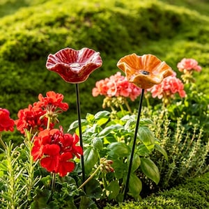 Könnte beinhalten: Zwei Gartenstecker aus Keramik in Blumenform, einer in tiefem Rot, der andere in Orange. Beide haben ein strukturiertes, blütenblattartiges Design. Die Stecker stehen in einem Garten mit roten und rosa Blumen, grünen Pflanzen und einem moosigen Hintergrund.