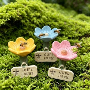 Könnte beinhalten: Drei blütenförmige Bienenwasserbecher in Gelb, Blau und Rosa, jeweils mit einer Biene obenauf. Jede Blume hat ein kleines Schild mit der Aufschrift "CUPS FOR BEE". Die Becher stehen in einem Bett aus grünem Moos.