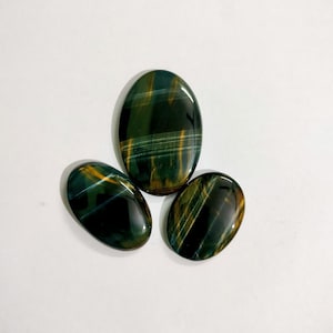 May include: Three polished gemstones with an oval shape. The stones have a dark green base color with streaks of gold and blue. The gemstones are arranged on a white surface.