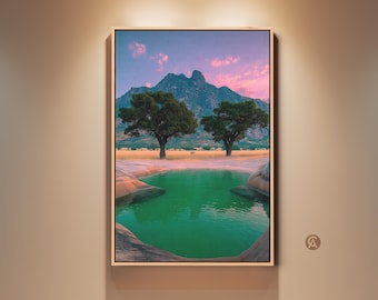 May include: A framed landscape art print featuring a serene scene with a green pond, two trees, and a mountain range under a pink and blue sky. The artwork is displayed on a beige wall.