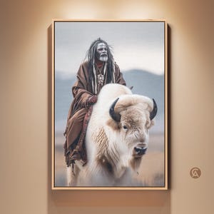 Peut inclure: Œuvre encadrée représentant une personne chevauchant un bison blanc. La personne porte une robe marron, avec du maquillage blanc sur le visage et de longs cheveux gris. Le bison a de grandes cornes noires, sur un paysage flou.