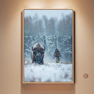 Può includere: Una stampa incorniciata raffigura un bisonte in un campo innevato, rivolto verso lo spettatore. Una persona con un fucile cammina sullo sfondo. L'opera d'arte è ambientata su uno sfondo di alberi innevati, creando una scena invernale.