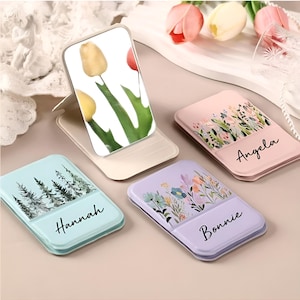Puede incluir: Un conjunto de soportes para teléfono personalizados en varios colores pastel. Cada soporte presenta un diseño floral o una escena de la naturaleza, con nombres como "Angela", "Hannah" y "Bonnie". Un soporte muestra una imagen de tulipán.