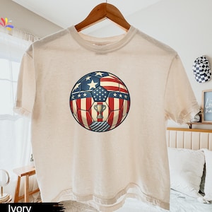 May include: An ivory short-sleeve t-shirt featuring a central graphic of a football patterned with the American flag design, including red and white stripes and white stars on a blue field, with a small gold trophy icon centred on the ball.