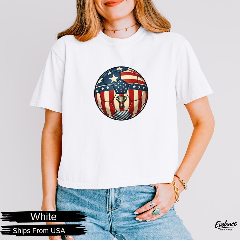 May include: A white short-sleeved t-shirt featuring a central graphic of a football patterned with the American flag design, including stars and stripes, and a small gold trophy icon centred within the ball's paneling.