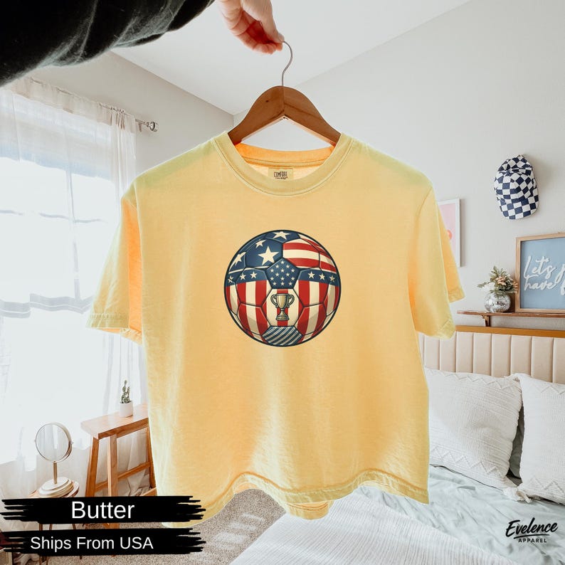 May include: A pale yellow short-sleeve t-shirt featuring a central graphic of a football patterned with the American flag design, including white stars on a blue field and red and white stripes, with a small gold trophy icon in the centre.