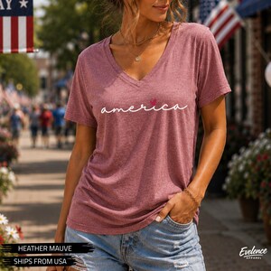Peut inclure: Un t-shirt &agrave; manches courtes de couleur mauve chin&eacute; avec une encolure en V. Le devant du t-shirt pr&eacute;sente le mot "america" imprim&eacute; en &eacute;criture cursive blanche, avec un petit c&oelig;ur rouge rempla&ccedil;ant le point sur la lettre "i". Le tissu est textur&eacute;.