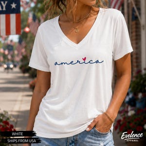 Peut inclure: Un t-shirt blanc &agrave; manches courtes et col en V avec le mot "america" &eacute;crit en lettres cursives bleu fonc&eacute; sur la poitrine, accompagn&eacute; d'un petit c&oelig;ur rouge plac&eacute; au-dessus de la lettre "i" au centre du motif graphique.