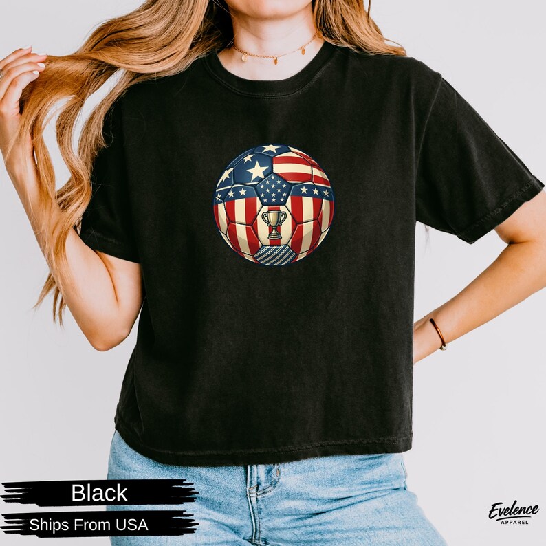 May include: A black short-sleeved t-shirt featuring a central graphic of a football patterned with the red, white, and blue colours of the American flag, including stars and stripes, with a small gold trophy icon centred on the ball.