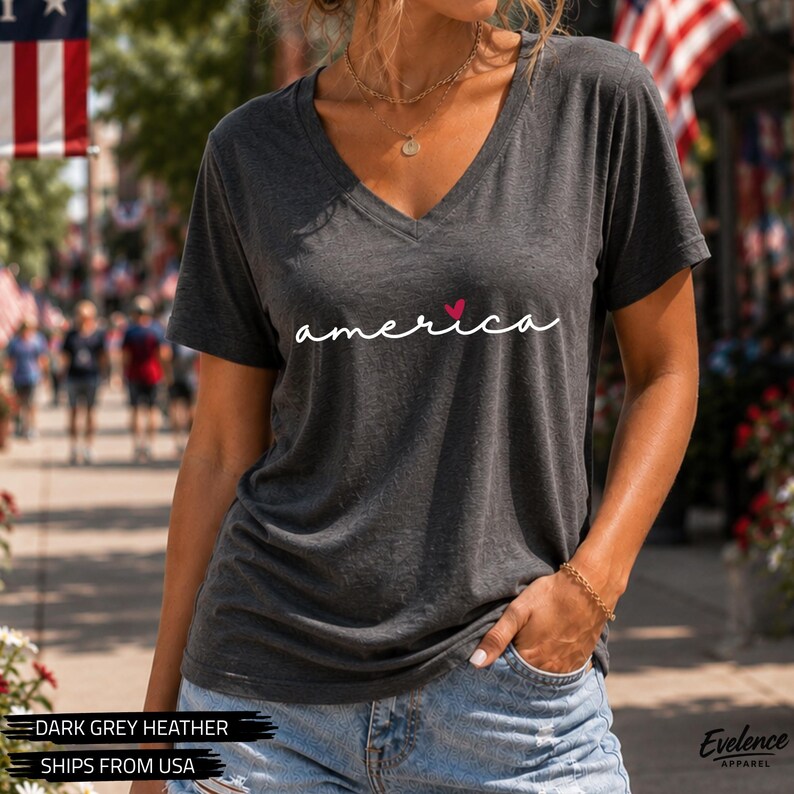 Peut inclure: Un t-shirt &agrave; col en V gris anthracite chin&eacute; avec le mot america &eacute;crit en lettres cursives blanches, dot&eacute; d'un petit c&oelig;ur rouge rempla&ccedil;ant le point sur la lettre i. Le v&ecirc;tement pr&eacute;sente des manches courtes et une coupe d&eacute;contract&eacute;e.