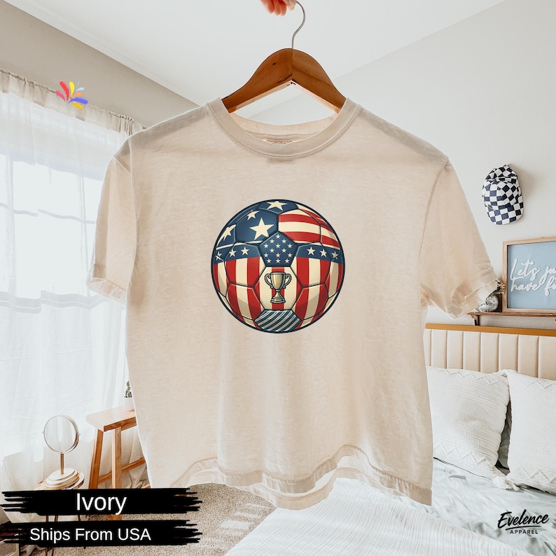 May include: An ivory short-sleeve t-shirt featuring a central graphic of a football patterned with the American flag design, including red and white stripes and white stars on a blue field, with a small gold trophy icon centred on the ball.