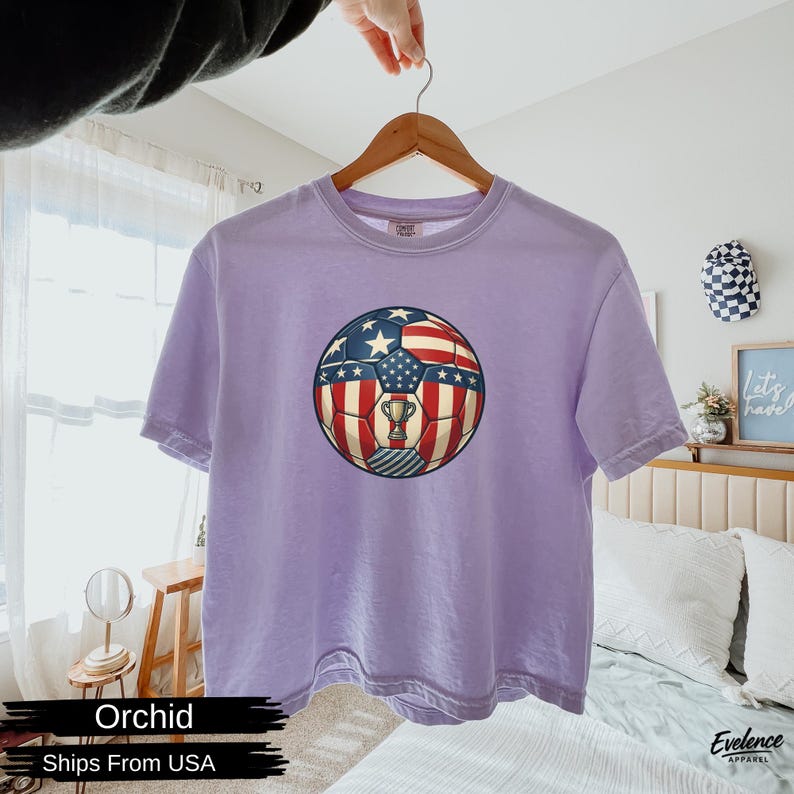 May include: A light purple short-sleeve t-shirt featuring a central graphic of a football patterned with the American flag design, including red and white stripes, blue sections with white stars, and a small silver trophy icon in the centre.