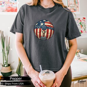 May include: A charcoal grey short-sleeved t-shirt featuring a central graphic of a football patterned with the American flag design, including red and white stripes and blue sections with white stars, and a small gold trophy icon in the centre.