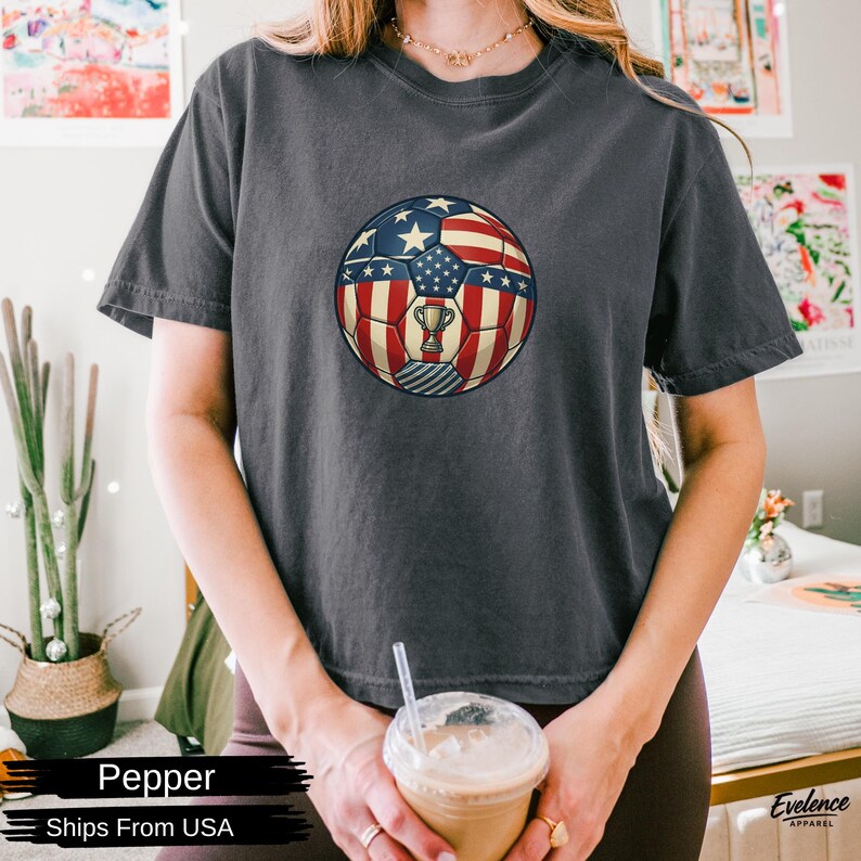 May include: A charcoal grey short-sleeved t-shirt featuring a central graphic of a football patterned with the American flag design, including red and white stripes and blue sections with white stars, and a small gold trophy icon in the centre.