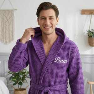 May include: A purple waffle-knit bathrobe featuring a hood, long sleeves, and a matching tie belt at the waist. The left chest area is embroidered with the name Liam in white cursive script. The fabric has a distinct grid-like textured pattern.