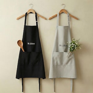 May include: Two aprons hanging on wooden hangers. One is black with "Mr. Eriksen" printed on it and a wooden spoon in the pocket. The other is gray with "Mrs. Eriksen" printed on it and a small bouquet of white flowers.