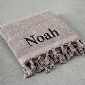 以下が含まれることがあります： 黒で「Noah」と刺繍された、折り畳まれたトープ色のタオル。 タオルはフリンジエッジで、柔らかく質感のある素材で作られています。 タオルは灰色の表面にきちんと折りたたまれています。