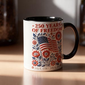 Puede incluir: Taza de cerámica blanca con asa e interior negros. La taza presenta un diseño con el texto "*250 YEARS OF FREEDOM" y una bandera estadounidense rodeada de elementos florales rojos y azules. La parte inferior de la taza dice "July 4, 1776 - July 4, 2026".