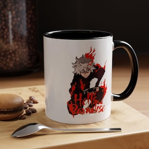 May include: White ceramic mug with a black interior and handle. The mug features a red and black anime-style illustration of a character with the text "Hell's Paradise". A spoon and a macaron are on a wooden surface.