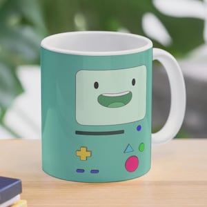 Puede incluir: Una taza de cerámica blanca con un asa negra y el texto "But First, Coffee" en negrita negra. La taza está sobre un fondo borroso. La taza mide aproximadamente 10 cm de altura.