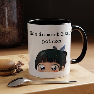 Puede incluir: Taza de cerámica blanca con interior y asa negros. La taza presenta el texto "This is most likely poison" sobre una ilustración de estilo anime de un personaje con ojos grandes. Una cuchara y un macaron están sobre la superficie de madera.