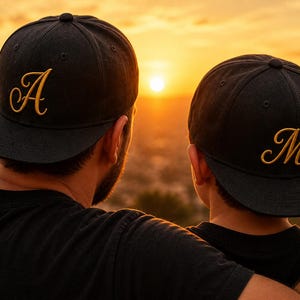 Könnte beinhalten: Zwei schwarze Baseballkappen mit goldgestickten Buchstaben, "A" und "M", sind zu sehen. Die Kappen werden von zwei Personen getragen, von hinten gesehen, mit einem orangefarbenen Sonnenuntergang im Hintergrund. Die Kappen haben einen gebogenen Schirm.