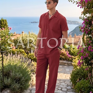 May include: A brick-red linen shirt and matching pants set. The short-sleeved shirt has a button-down front and a chest pocket. The pants are straight-legged. The outfit is worn with sunglasses and sandals. The word "SARTORIA" is partially visible.