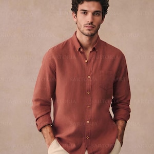 May include: A rust-colored button-down shirt with a chest pocket and long sleeves. The shirt is paired with light beige pants. The shirt has a classic collar and gold-colored buttons. The background is a neutral beige.