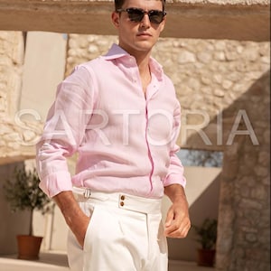 Puede incluir: Una persona con una camisa rosa claro abotonada y pantalones blancos. La camisa tiene mangas largas y cuello clásico. Los pantalones tienen cintura alta y corte entallado. La persona también lleva gafas de sol. La palabra "SARTORIA" es visible en la camisa.