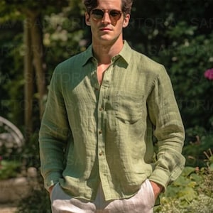 May include: A man wearing a light green linen button-down shirt with a chest pocket and beige pants. He is also wearing sunglasses. The shirt is unbuttoned at the top, and he has his hands in his pockets. The background is a blurred outdoor scene.