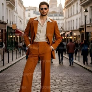 Puede incluir: Un hombre con un traje color óxido con pantalones de campana y una camisa color crema. La chaqueta del traje tiene solapas anchas. El hombre lleva gafas de sol y está de pie en una calle adoquinada. El fondo presenta edificios y personas.