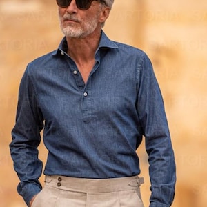 May include: A man wearing a blue denim long-sleeve shirt with a Henley collar and beige pleated trousers. The shirt has a buttoned placket and the trousers have a high waist. The man is also wearing sunglasses.